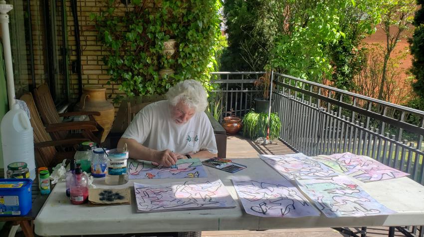 Artista dibujando rodeado de bocetos sobre una mesa en el balcón de su casa.