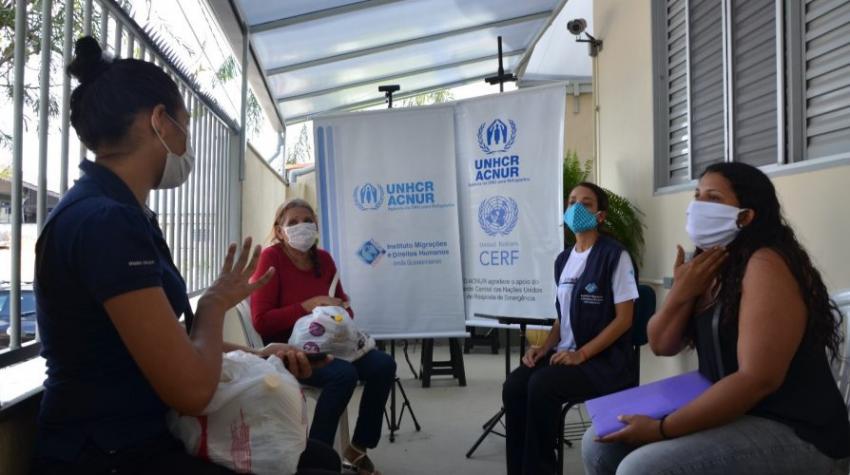 A program officer speaks to Francis, Silany and Silany's mother to issue them a benefit card in Brasilia. All four of them are wearing facemasks. They are seated in a courtyard outside a building.