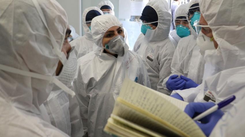 WHO and public health experts are deep in discussion whilst one writes notes. They are dressed in head-to-toe protective gear including face masks.