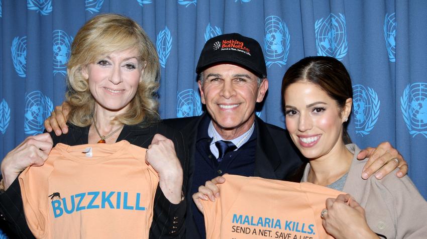 Two women and one man holding orange t-shirts saying "Malaria Kills. Send a net. Save a light." 