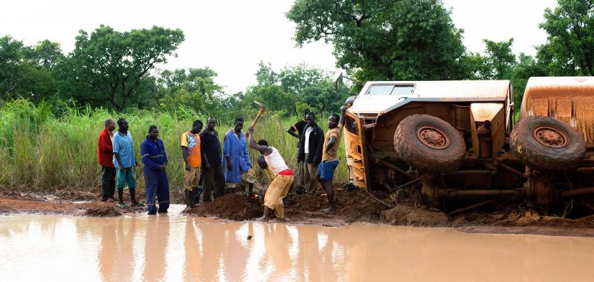 A group of truck drivers take turns clearing earth to drain water from an impassable section of road and an overturned truck is behind them.