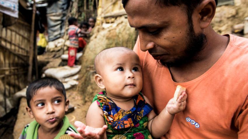 Kamal cradles his daughter who is holding a piece of bread, while a boy fondly looks at them.