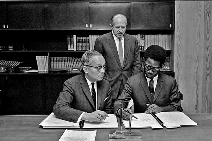 UN Ambassador signing Convention with UN Secretary-General at left (both seated) and UN Legal Counsel standing above them.
