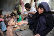 A mother wearing a black abayia feeds a child some rice by hand. The child is in a disheveled state and is sparsely dressed. In the background are more children and a man.