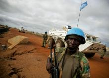 Zambian peacekeeper is standing in front of white tank with the UN emblem and the flag. 