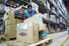 Man wearing a face-mask and gloves sorts through boxes in a warehouse.