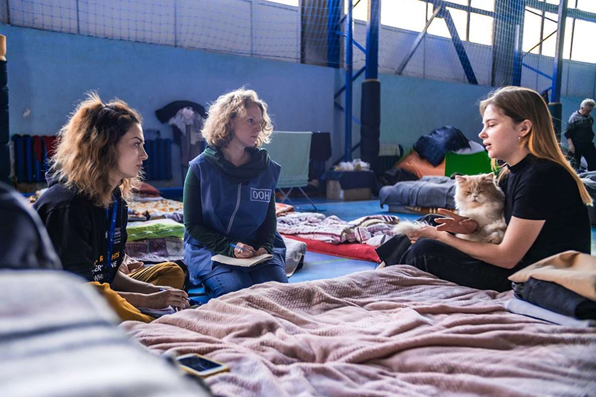 Two human rights officers sit on cots listening to a woman, who has a cat on her lap.