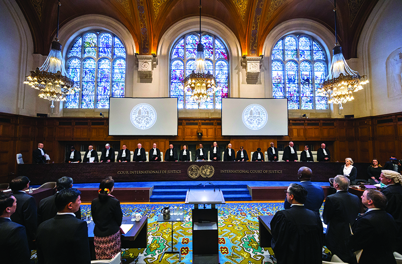 Panoramic view of a courtroom with two ornate chandeliers and stained-glass windows. Seventeen judges sit behind a long podium against the far wall. Two screens hang on the wall behind them. A group of people sit at tables facing the judges and the screens.