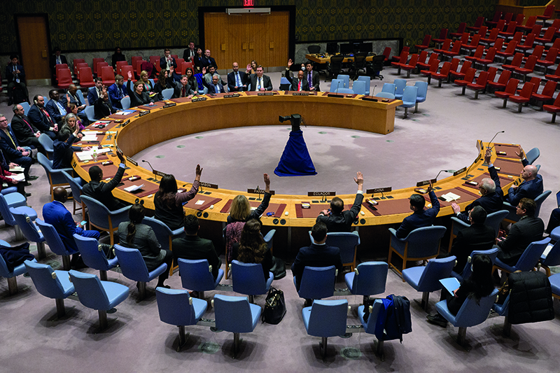 A group of people are seated around a circular table in a meeting room. Many of them have their hands raised. A projector stands in the middle of the room.