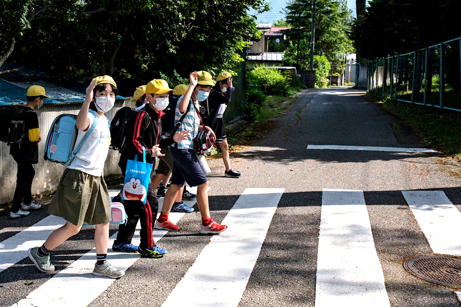 Group of children wearing facemasks wave as they walk through a crosswalk.  