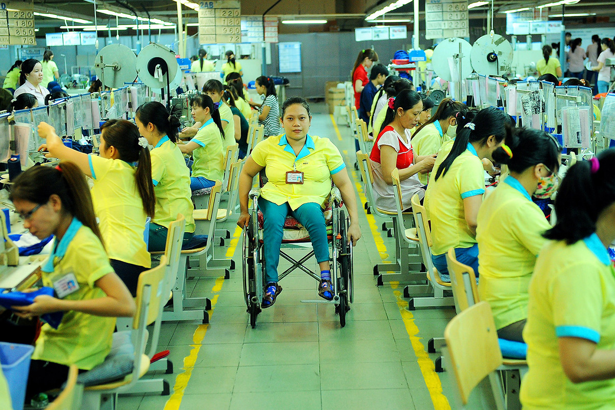 A factory worker in a wheel goes down the hall among women at sewing machines.