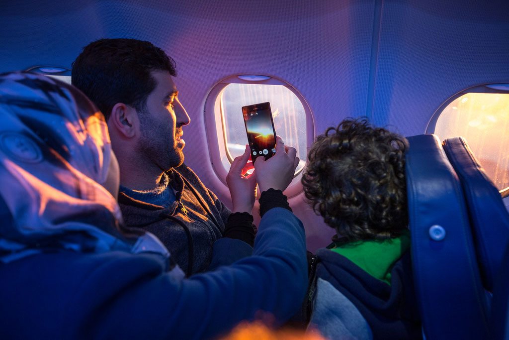 Una familia mira por la ventana del avión y toma fotos mientras viajan a su nuevo hogar. Foto: OIM / Muse Mohammed