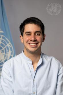 ASG Felipe Paulier, a smiling man with brown hair and brown eyes, stands in front of the United Nations flag.