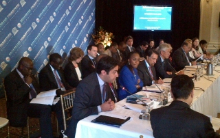 Man sitting at long table speaking into microphone with a woman and 4 other men seated next to him