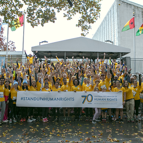 在“冲向终点”5公里长跑比赛前，参赛的联合国工作人员手持“挺身维护人权”的横幅在总部门前合影，摄于2018年11月3日。联合国图片